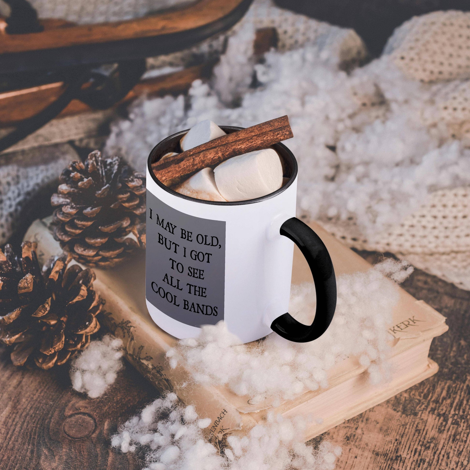 White mug with black handle and text, surrounded by cinnamon sticks and pine cones on a textured surface.
